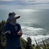 Person wearing a cap and sunglasses standing on a coastal cliff with ocean and sky in the background